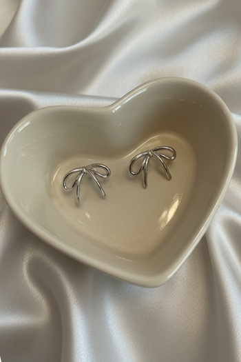 Small silver bow-shaped stud earrings with a fine ribbon detail on a soft fabric background.