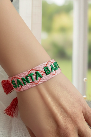 A pink woven friendship bracelet with "SANTA BABY" embroidered in green letters, accented with red diamond outlines and pink tassels.