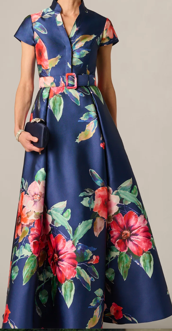 A woman models a navy blue satin-like maxi dress with a vibrant red, pink, and green floral print. The dress features short cap sleeves, a V-neckline, a matching fabric belt with a gold-tone buckle, and is styled with a small dark clutch.