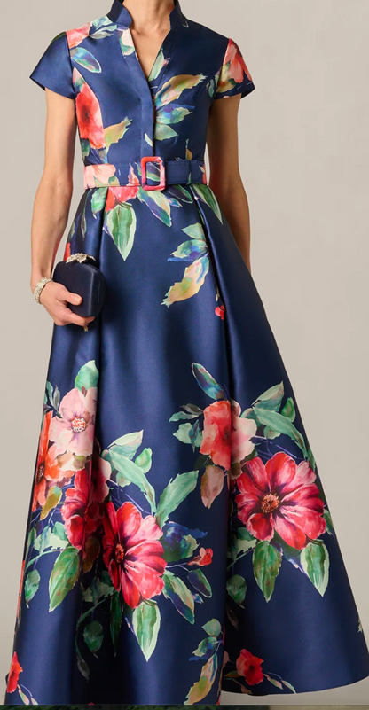 A woman models a navy blue satin-like maxi dress with a vibrant red, pink, and green floral print. The dress features short cap sleeves, a V-neckline, a matching fabric belt with a gold-tone buckle, and is styled with a small dark clutch.