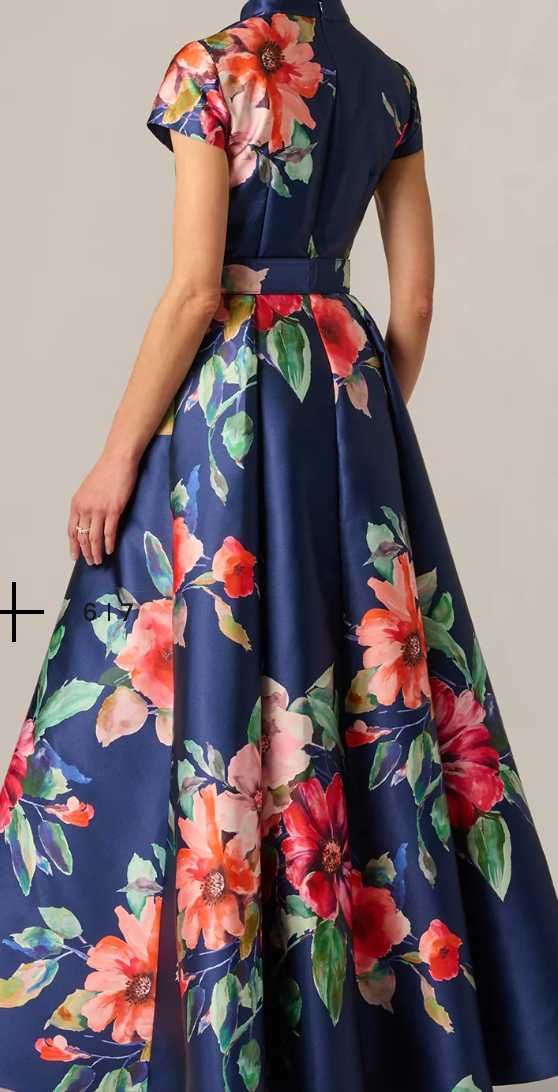 A woman shown from the back wearing a navy blue satin-like maxi dress with a vibrant floral print in red, pink, orange, and green. The dress features short sleeves, a structured high neckline, a matching waist belt, and a full flared skirt.