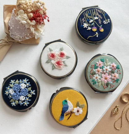 Set of compact mirrors with floral and nature-themed embroidery on a white surface.