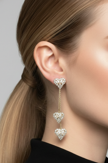 A pair of gold-toned drop earrings featuring three graduated, diamond-encrusted heart charms suspended from delicate chains, presented on a minimalist grey background.