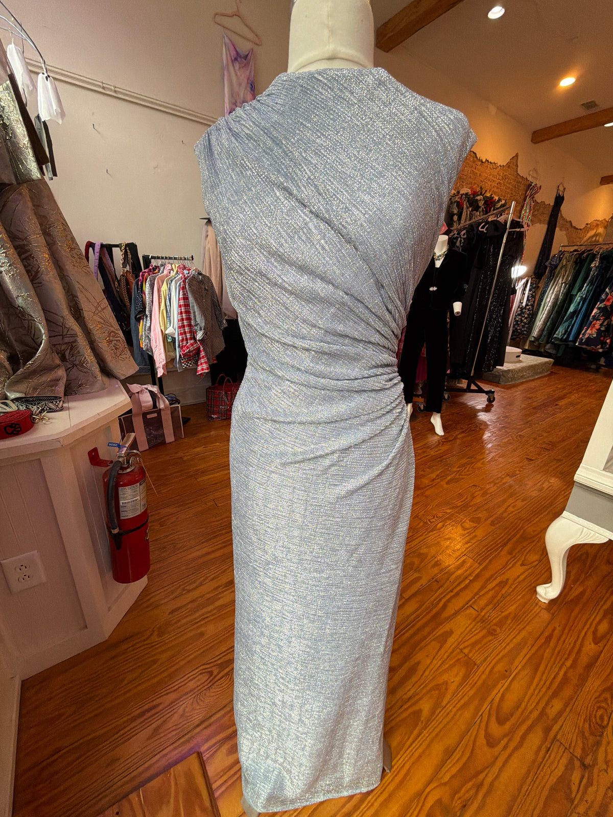 Periwinkle metallic gown with a ruched waist on a mannequin beside clothing racks in the boutique.