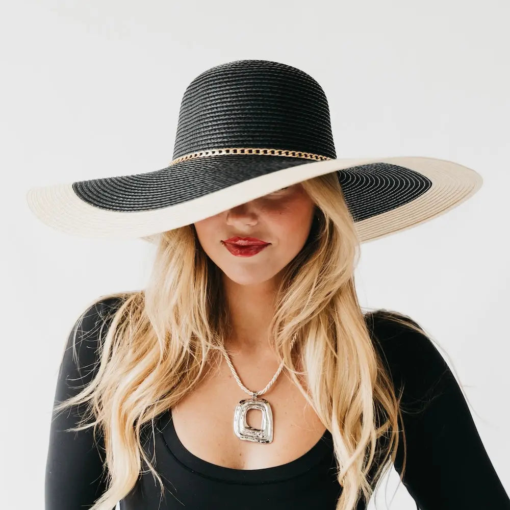 A blonde woman wearing a wide-brimmed black and beige straw sun hat with a gold chain detail.
