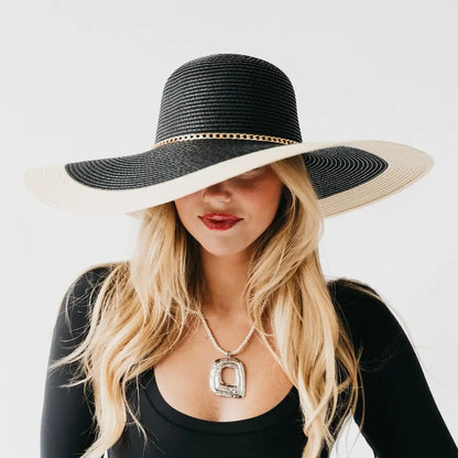 A blonde woman wearing a wide-brimmed black and beige straw sun hat with a gold chain detail.