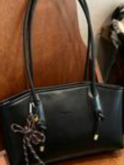 Elegant black women’s shoulder tote bag with long straps and decorative charm detail, displayed against a warm wood background.