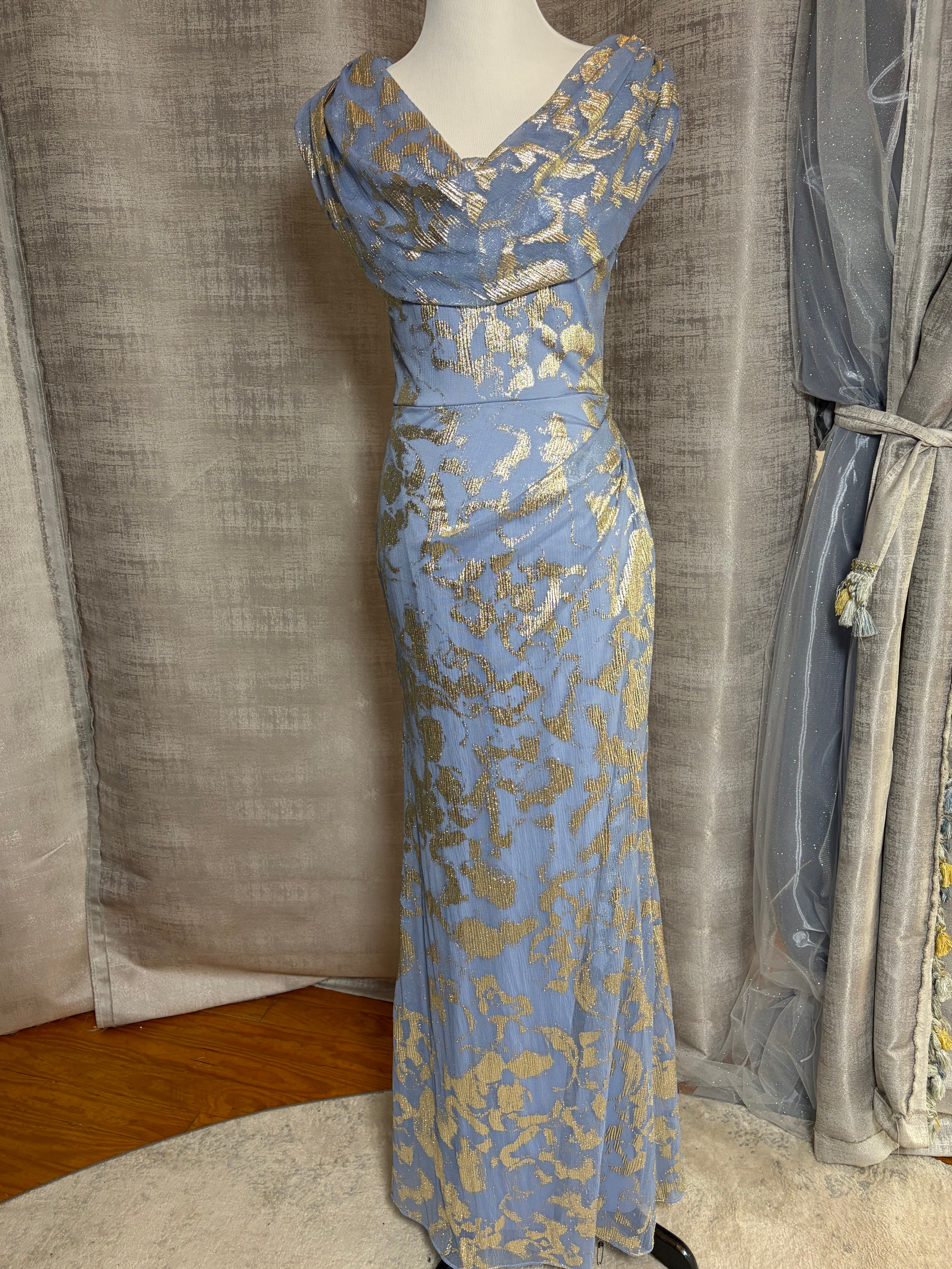 A floor-length light blue evening gown with a gold abstract floral pattern, displayed on a white mannequin in front of gray fabric curtains.