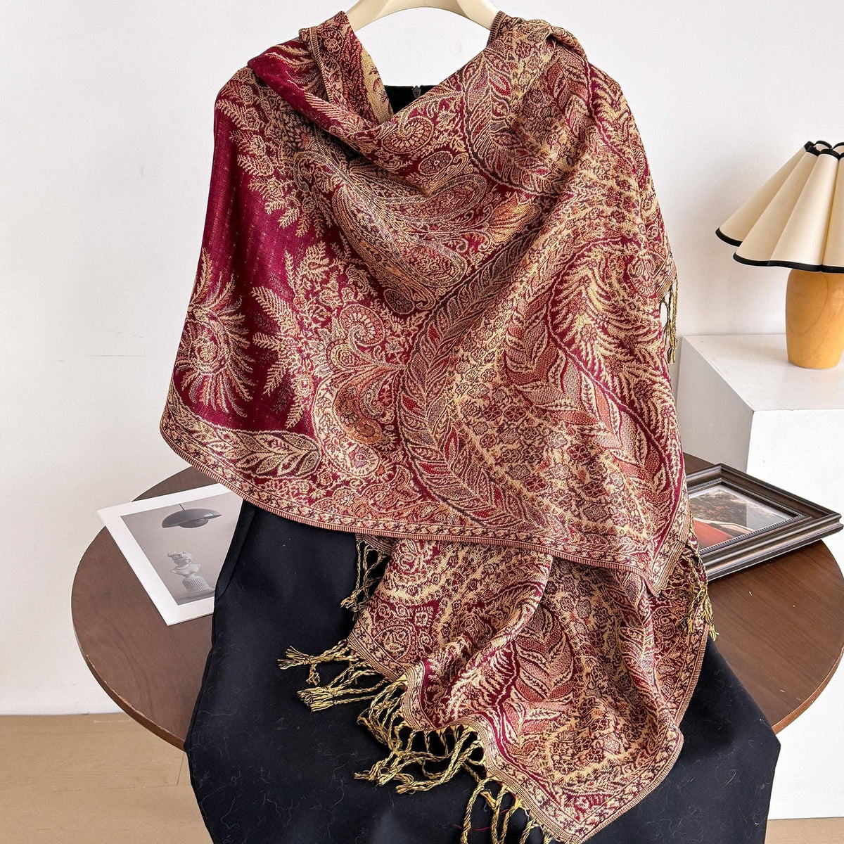 Red and gold patterned woven wrap draped over black fabric on a hanger, showing fringed ends.