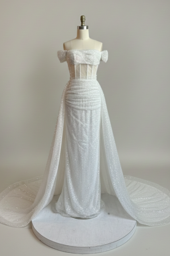 A shimmering white, sequin-covered gown featuring an off-the-shoulder neckline, a structured corset-style bodice with vertical boning, and a dramatic sheer overskirt extending into a side train.