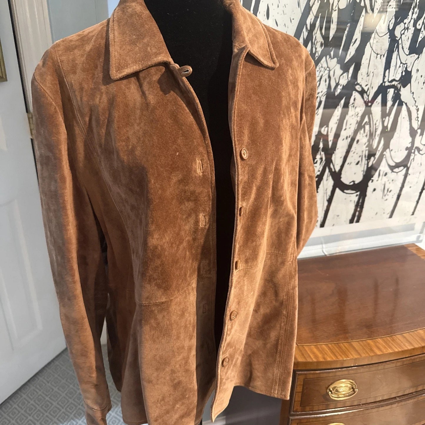 Brown suede jacket on a mannequin in front of a wooden dresser with a patterned curtain.