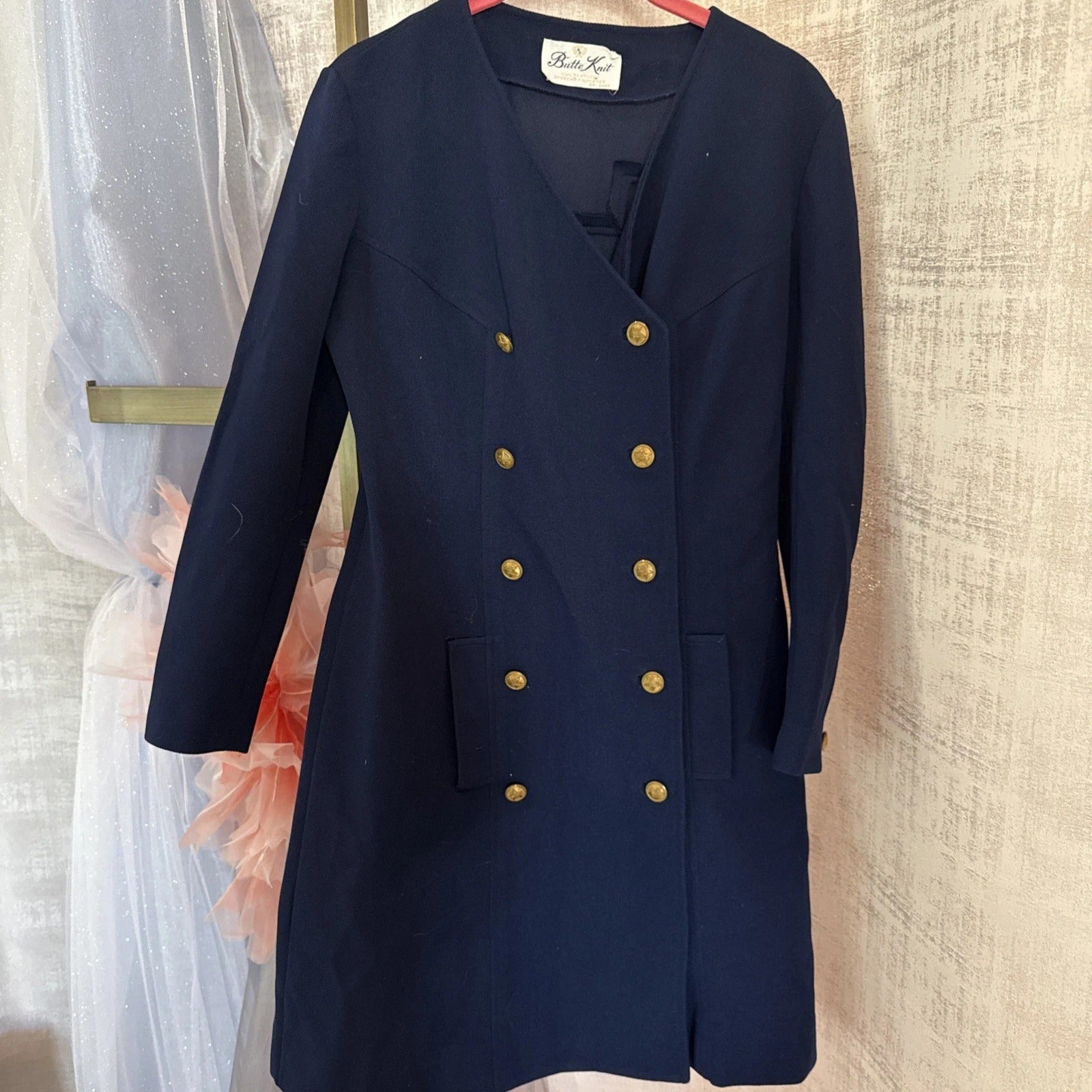 Navy blue coat with gold buttons on a hanger against a neutral wall.