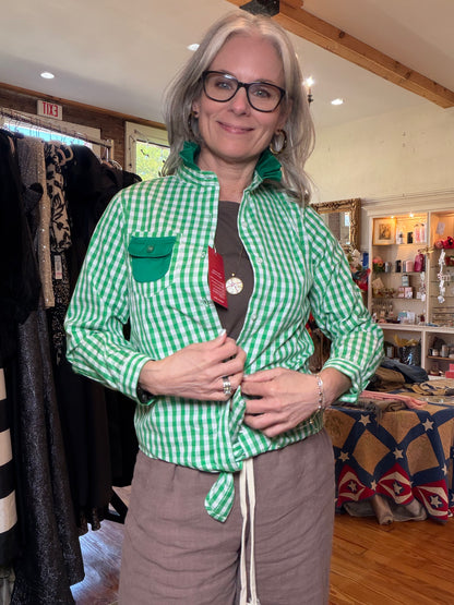 Front view of an open green gingham shirt with long sleeves, a ruffle collar and green pocket.